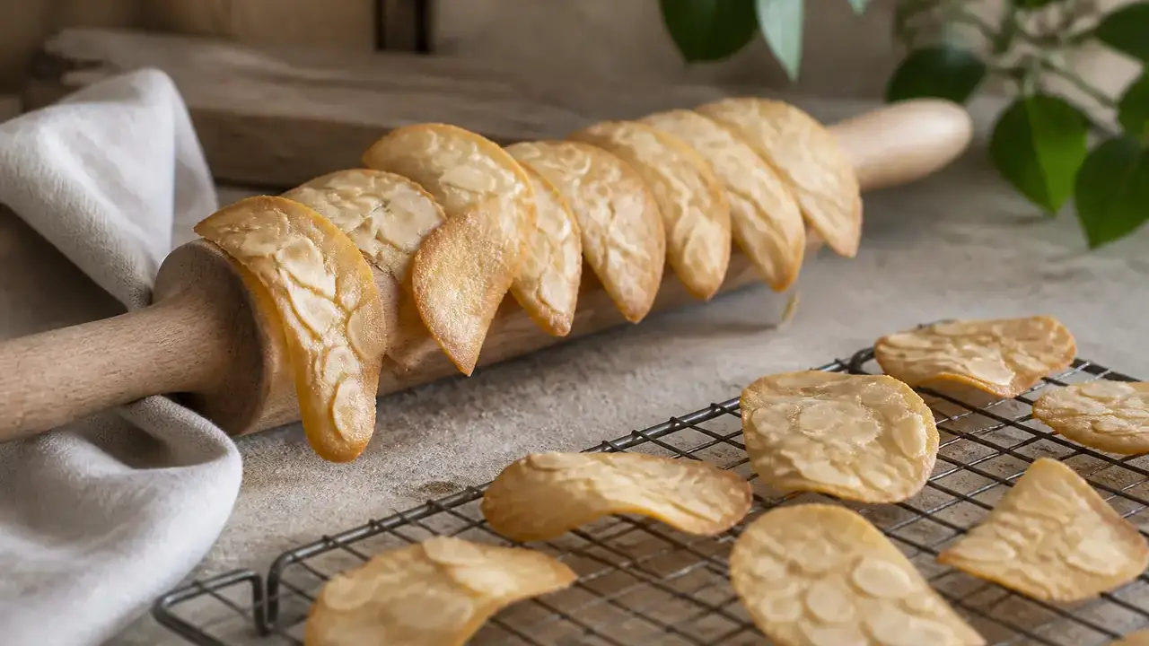 Tuiles aux amandes effilées, dorées sur les bords et incurvées, posées sur un rouleau à pâtisserie en bois et sur une grille