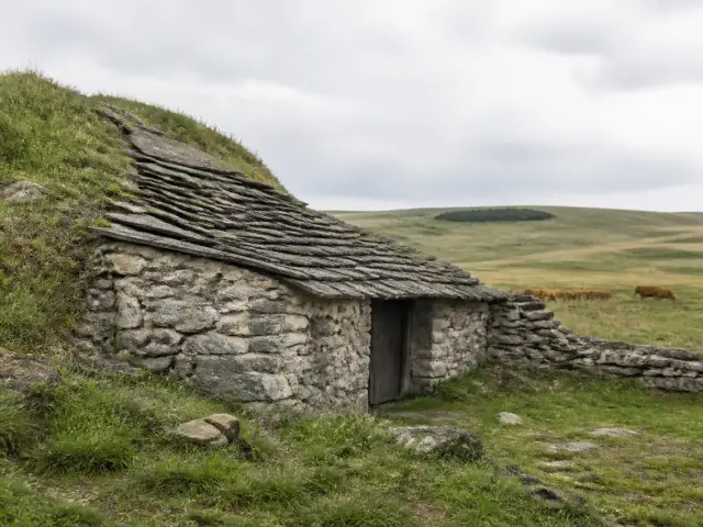 Une petite maison de pierre au toit de lauzes, adossée à une pente herbeuse, sur un plateau d’altitude