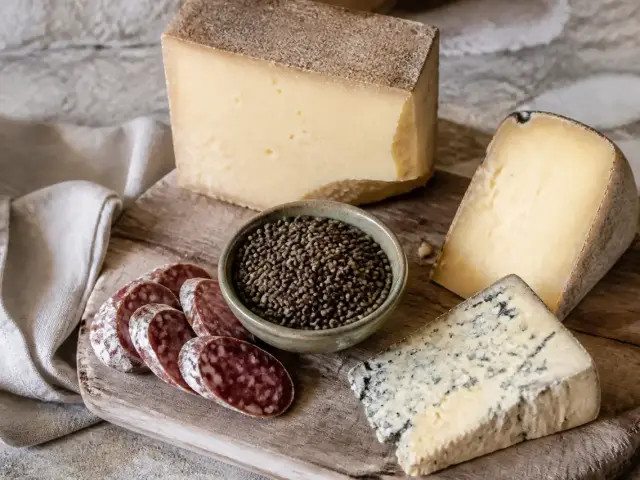 Une planche en bois avec plusieurs fromages de montagne entamés, un bol de lentilles vertes et quelques tranches de saucisson sec