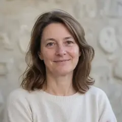 Portrait d’Héloïse Vernières, cheveux châtains mi-longs, pull de laine écru, sourire discret, devant un mur de pierre clair et flou