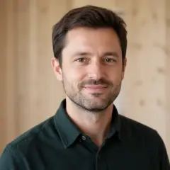 Portrait de Tristan Vigouroux, cheveux bruns courts, barbe de quelques jours, chemise en flanelle vert sombre, devant un fond de bois clair et flou